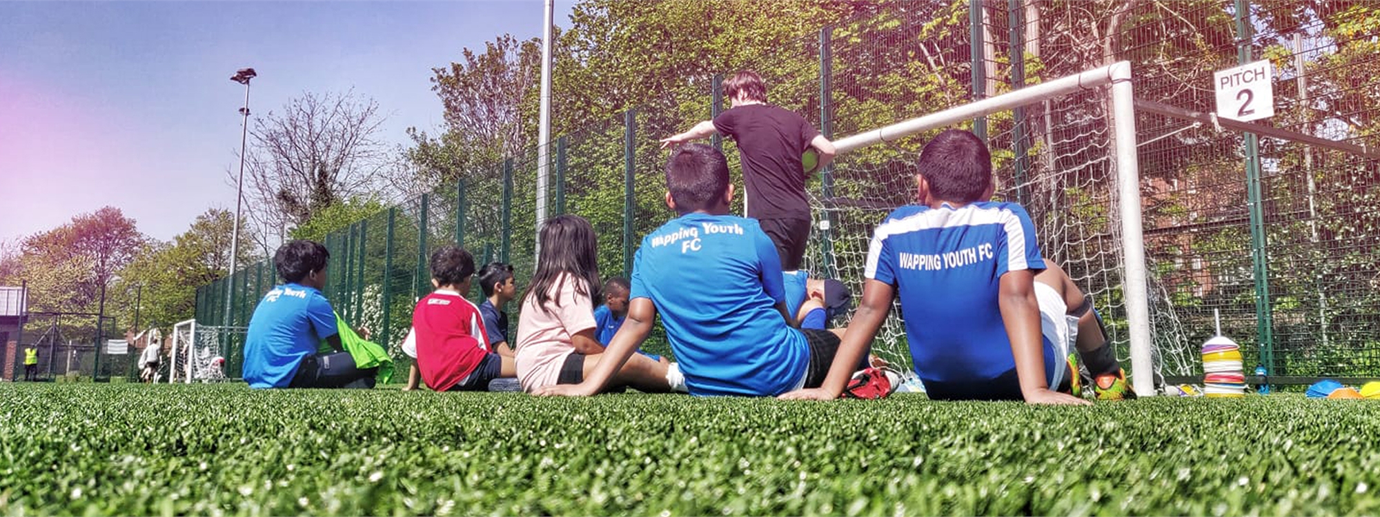 Kids sat on the floor in front of a football goal watching someone teaching